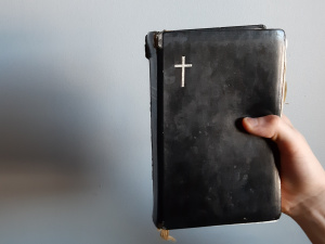 Kuvassa henkilö pitää kädessään Raamattua, jossa on mustat kannet ja kannessa kultainen risti. Tausta on vaaleansininen.